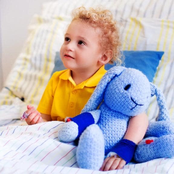 copil blod pe un pat de spital cu un tricou galben si un iepuras de plus albastru in brate
